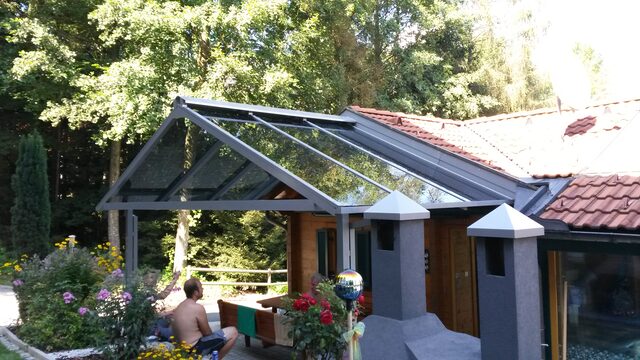 Verglaste Terrassenüberdachung mit Aluminium-Sparren an einem Holzhaus im Grünen