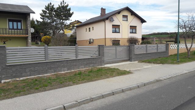Grauer Lamellenzaun mit Betonsockel und Doppelflügeltor vor einem Einfamilienhaus